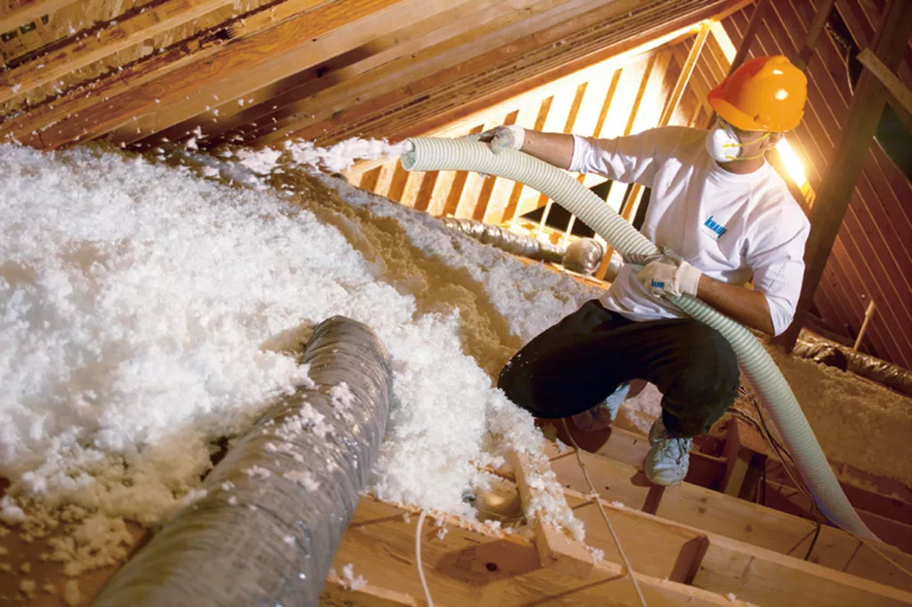 Technician wearing yellow hardhat while installing fiberglass blown insulation in an unfinished attic.